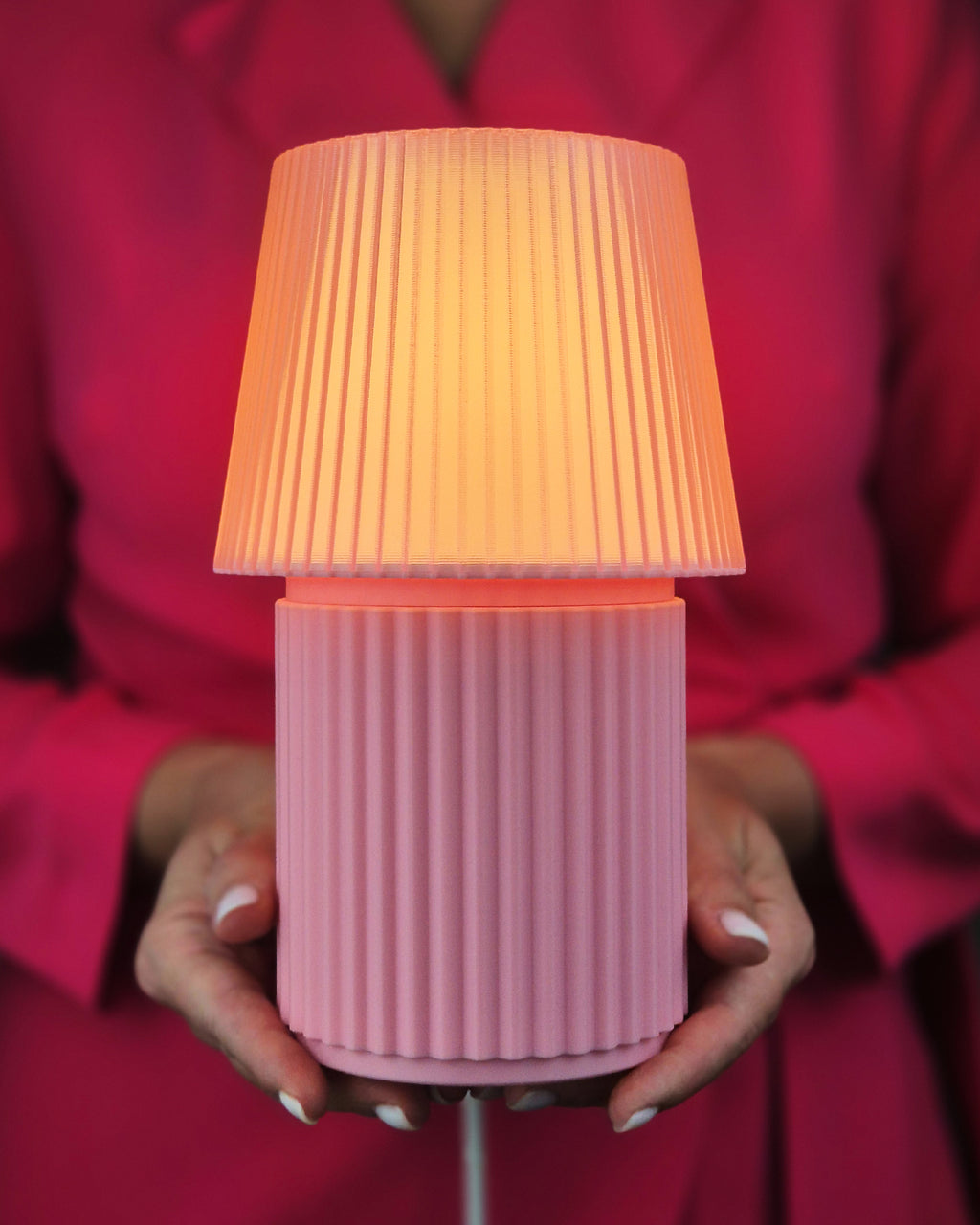 A pink ribbed table lamp with a pleated shade glowing warm orange, held by a person wearing a bright pink top.