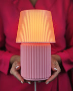A pink ribbed table lamp with a pleated shade glowing warm orange, held by a person wearing a bright pink top.