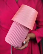 A close-up of a person holding a pink ribbed table lamp with a matching shade and base, wearing a bright pink blazer.