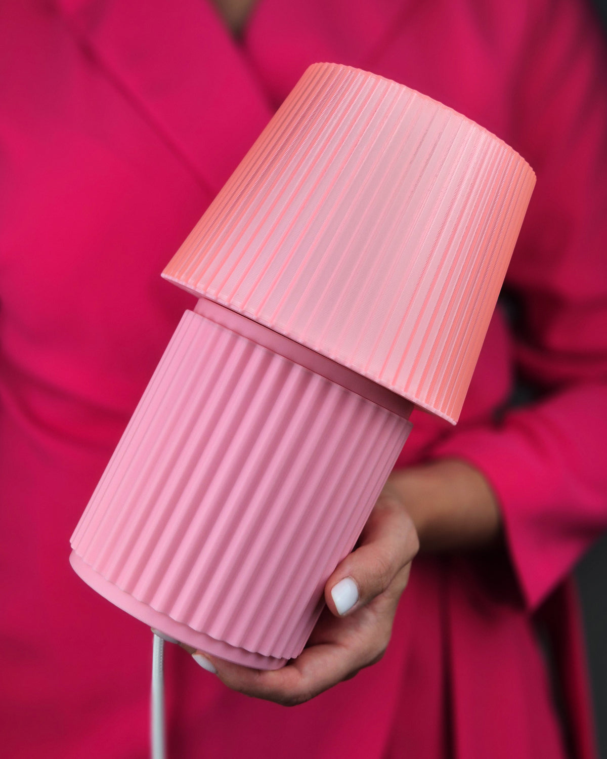 A close-up of a person holding a pink ribbed table lamp with a matching shade and base, wearing a bright pink blazer.