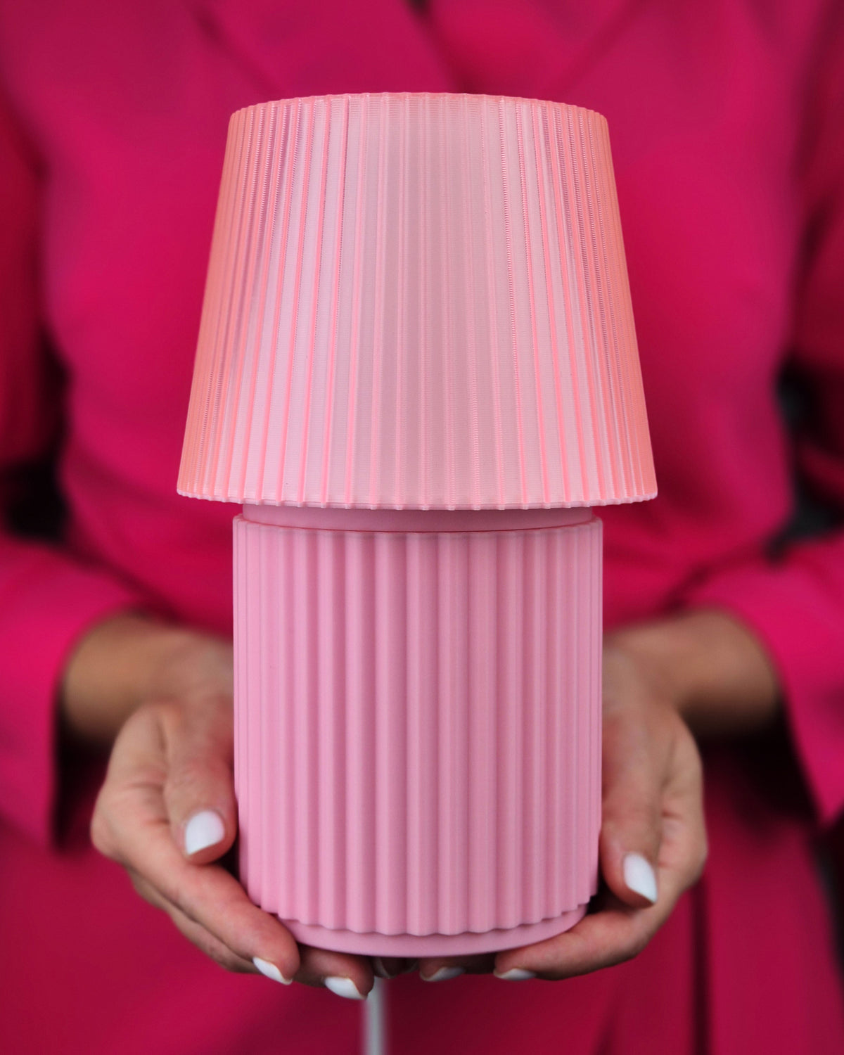 A pink pleated lampshade and matching ribbed cylindrical base held by hands against a bright pink blazer background.