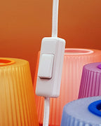 A close-up of a white corded switch with a rectangular toggle, attached to a white braided cord, set against a background of colorful ribbed plastic shades in orange, pink, purple, and blue.
